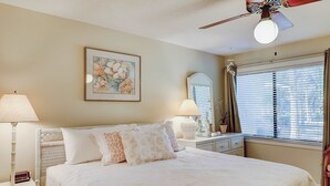 A neatly arranged bedroom with a bed, bedside lamps, a ceiling fan, a window with blinds, and a framed picture on the wall. - Terrific island escape with pool, furnished patio, fast WiFi & beach nearby (Hilton Head)