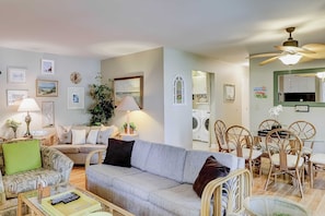 Ein Wohnzimmer mit einer Couch, Sesseln, einem Couchtisch, einem Essbereich mit Stühlen, einem Deckenventilator und einer Waschmaschine im Hintergrund.