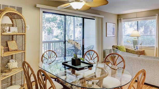 A dining area with a glass table, wicker chairs, a ceiling fan, and a view of the outdoors. - Terrific island escape with pool, furnished patio, fast WiFi & beach nearby (Hilton Head)