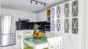 Une cuisine moderne avec des meubles blancs, un réfrigérateur en acier inoxydable, une table en verre avec des chaises et un panier à fruits.