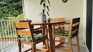 Une table et des chaises en bois, avec des coussins à motifs verts, sur un balcon doté d’une balustrade blanche et d’un pot de fleurs.