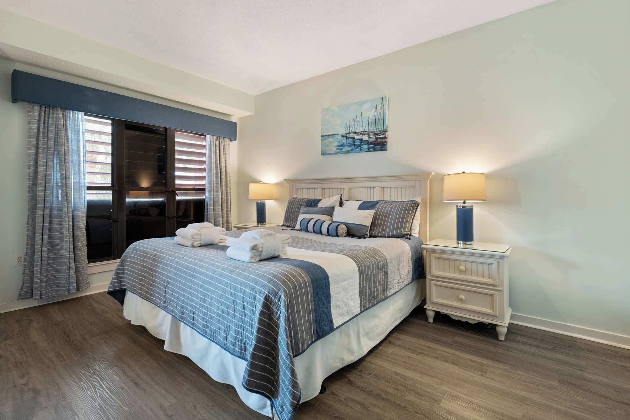 A bedroom with a bed, two bedside tables, a window with curtains, and a painting on the wall.
