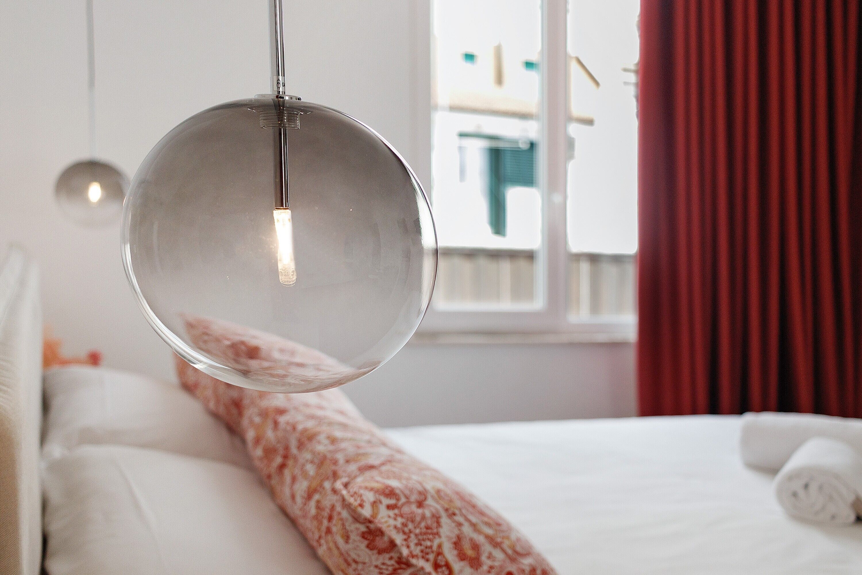 A modern bedroom with a hanging light fixture, a bed with white bedding, and a window with red curtains.