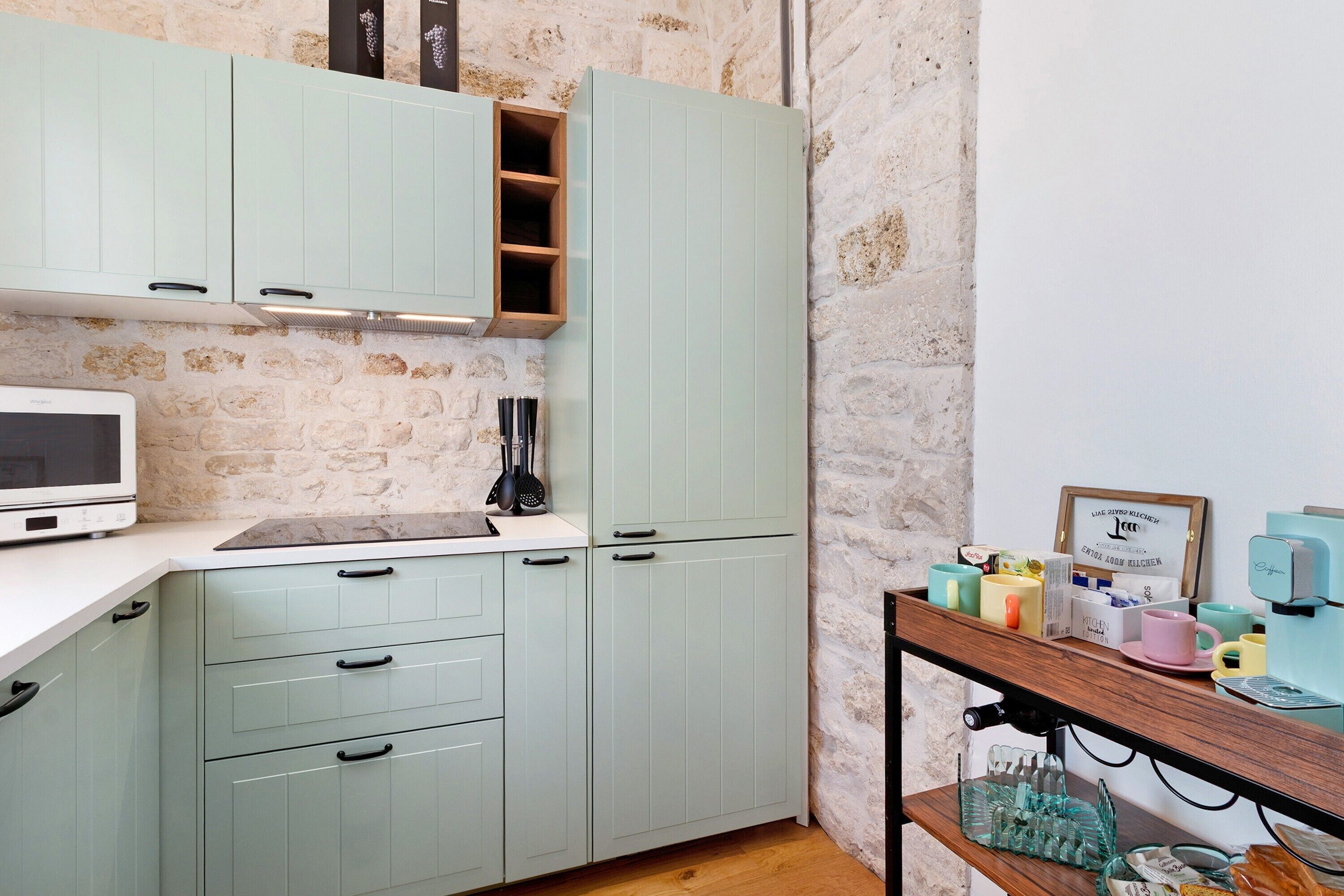 Una cocina con armarios verde claro, un microondas y una cafetera sobre una repisa de madera.