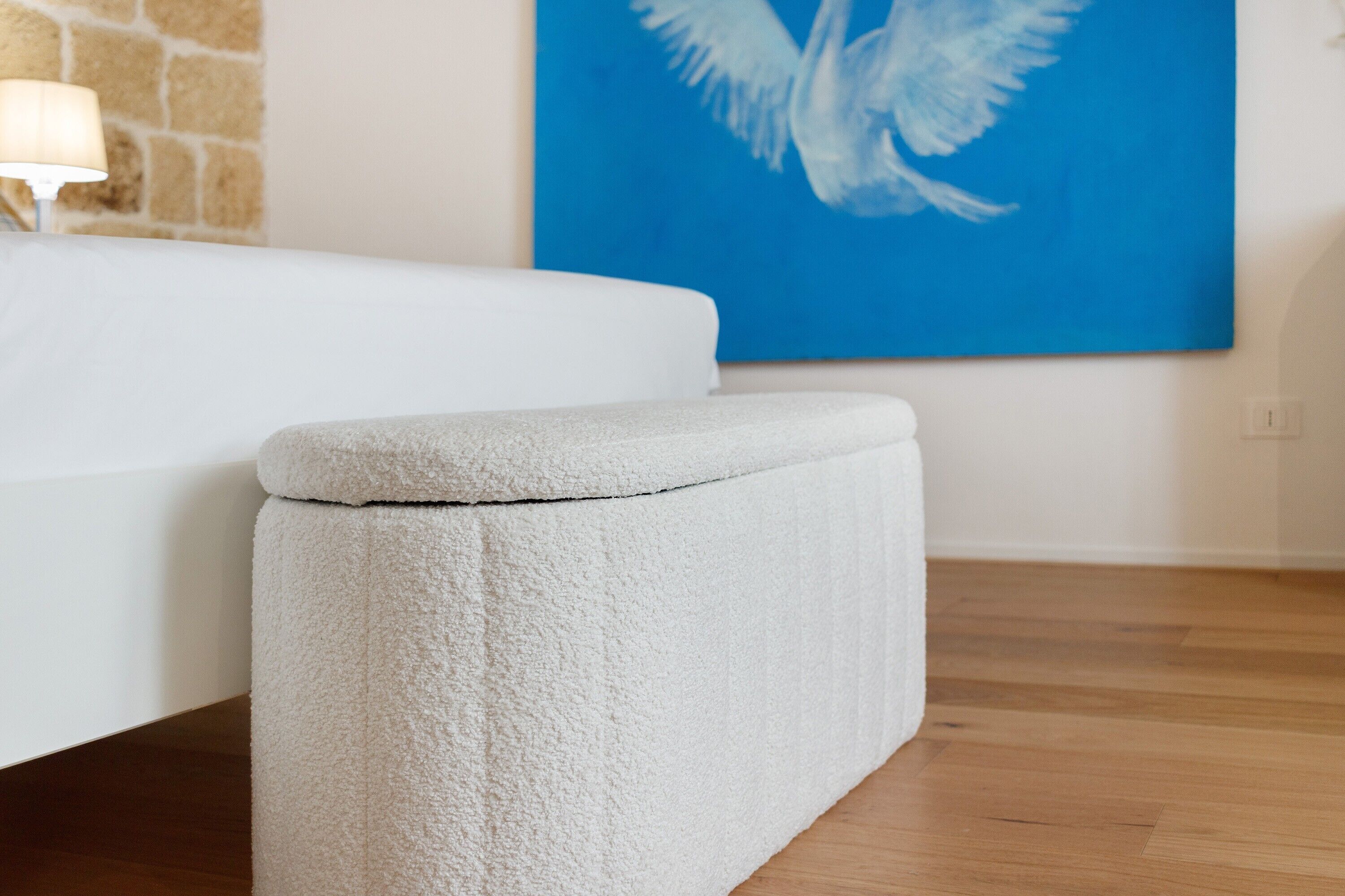 Un banc blanc dans une pièce dont le sol est en bois, avec un lit blanc et un mur bleu orné d’un tableau représentant un oiseau blanc.