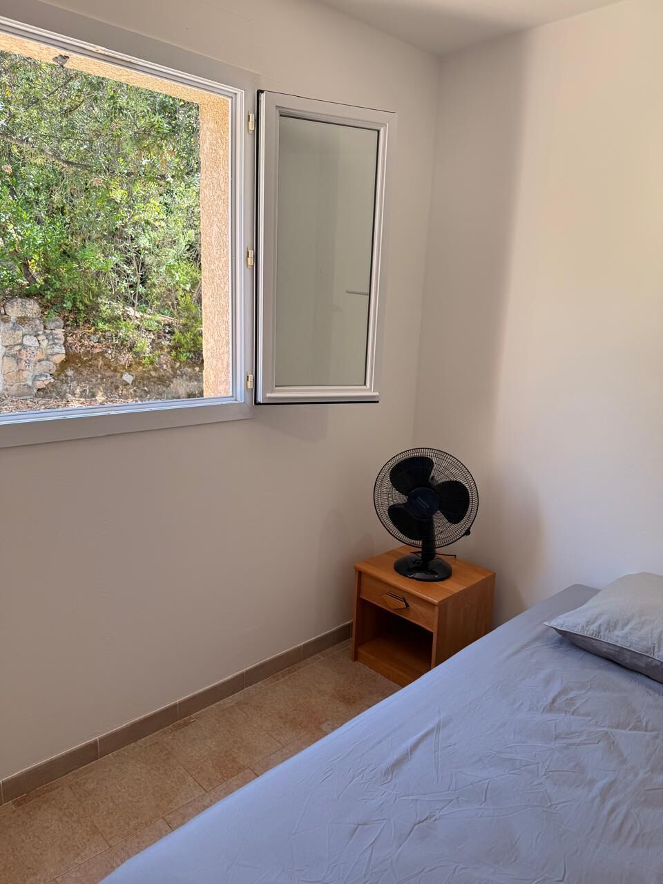 A bedroom with a bed, a window with a view of greenery, a standing fan, and a mirror on the wall.