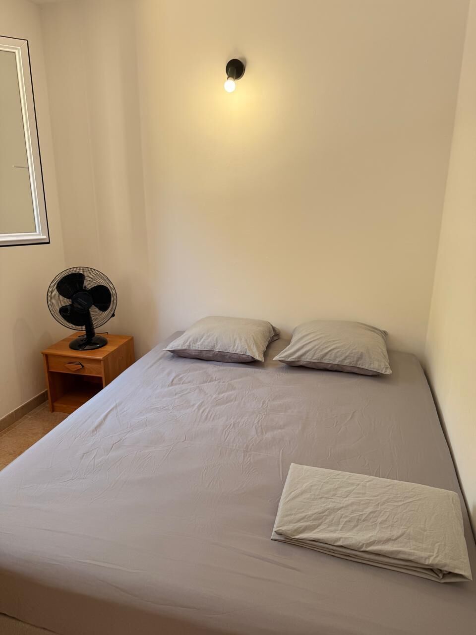 Una cama individual con dos almohadas, una mesita de noche con un ventilador y un aplique de pared.