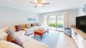 Een ruime woonkamer met plafondventilator, een grote flatscreen-tv, een zithoek, een salontafel en een schuifdeur naar een balkon.