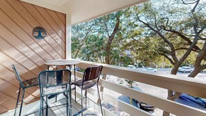 Een balkon met een tafel en stoelen, een decoratief object aan de muur, en uitzicht op bomen en geparkeerde auto's.