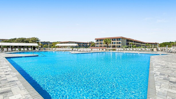 En stor swimmingpool med fliseterrasse, liggestole og en bygning med flere etager i baggrunden.