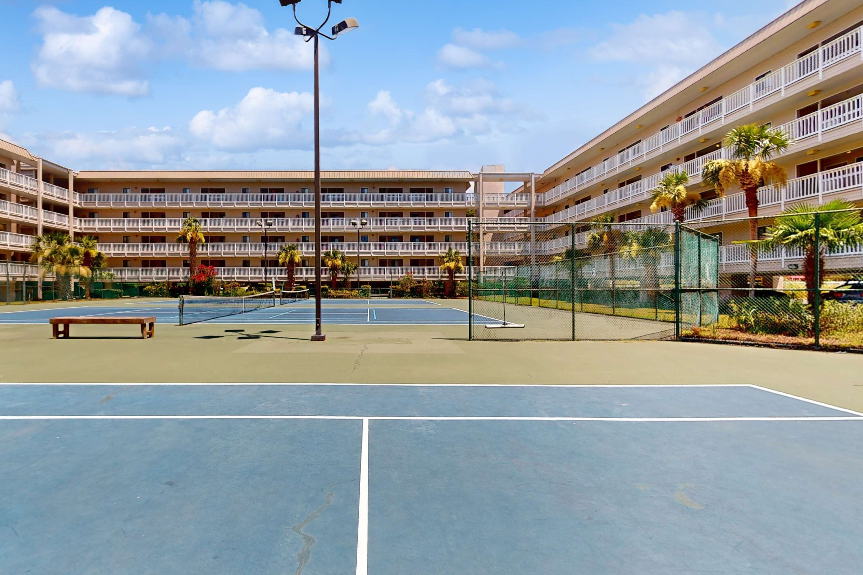 Un hôtel à plusieurs étages doté d’un court de tennis, de palmiers et d’un banc.