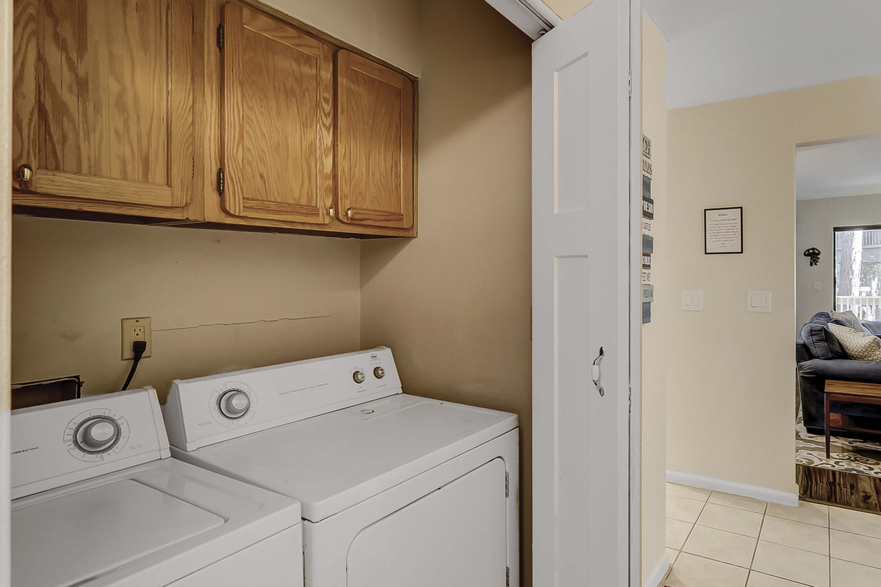 A laundry area with two white washers and dryers, wooden cabinets above, and a door leading to a living room with a couch.