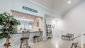 Une cuisine moderne avec une enseigne indiquant « MAISON DE PLAGE », un coin repas avec une longue table et des chaises, et un escalier avec une rampe.