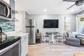 A modern kitchen with a built-in microwave, stainless steel refrigerator, and a coffee machine on a shelf. There is a dining area with a table and chairs, and a grey sofa with a pillow labeled 'Salt Sea Sand.' - Stunning Newly Renovated Condo with Ocean View & Shared Pool Close to the Beach (Hilton Head)