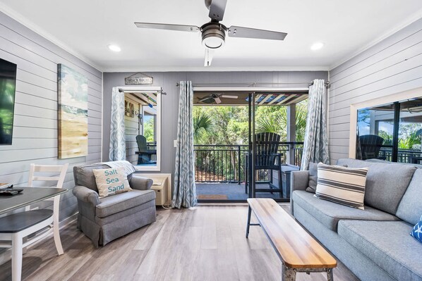 A modern living room with a ceiling fan, a sofa, a bench, a dining table, and a view of the outdoors. - Stunning Newly Renovated Condo with Ocean View & Shared Pool Close to the Beach (Hilton Head)
