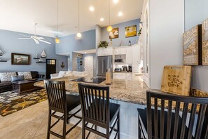 A modern kitchen with a central island, white cabinetry, and a dining area with a black table and chairs. - Nautical condo with a balcony, & AC - close to pickleball and tennis courts (Hilton Head)