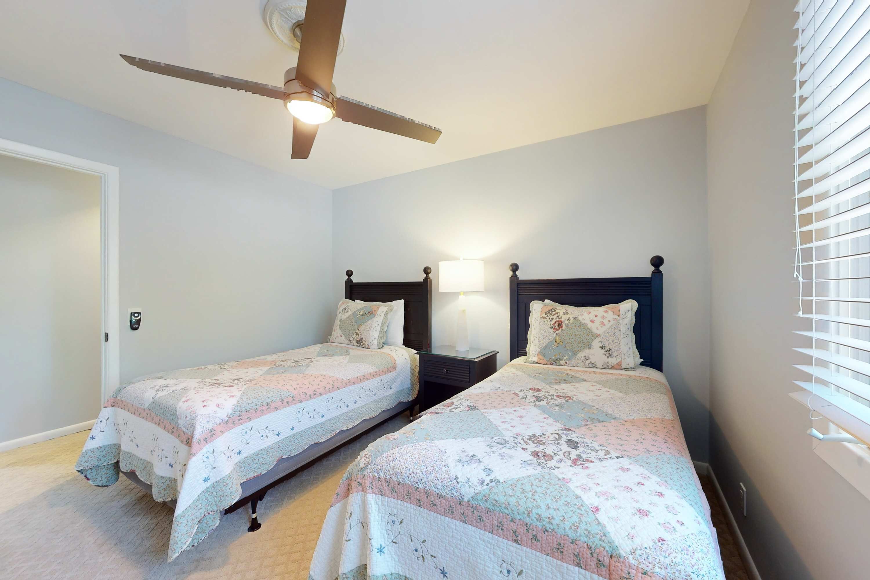 A bedroom with two beds, a ceiling fan, bedside lamps, and a window with blinds.