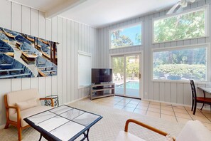 A living room with a TV, a sofa, a coffee table, and large windows. - Secluded home & pool house with private pool - steps away from the beach (Hilton Head)