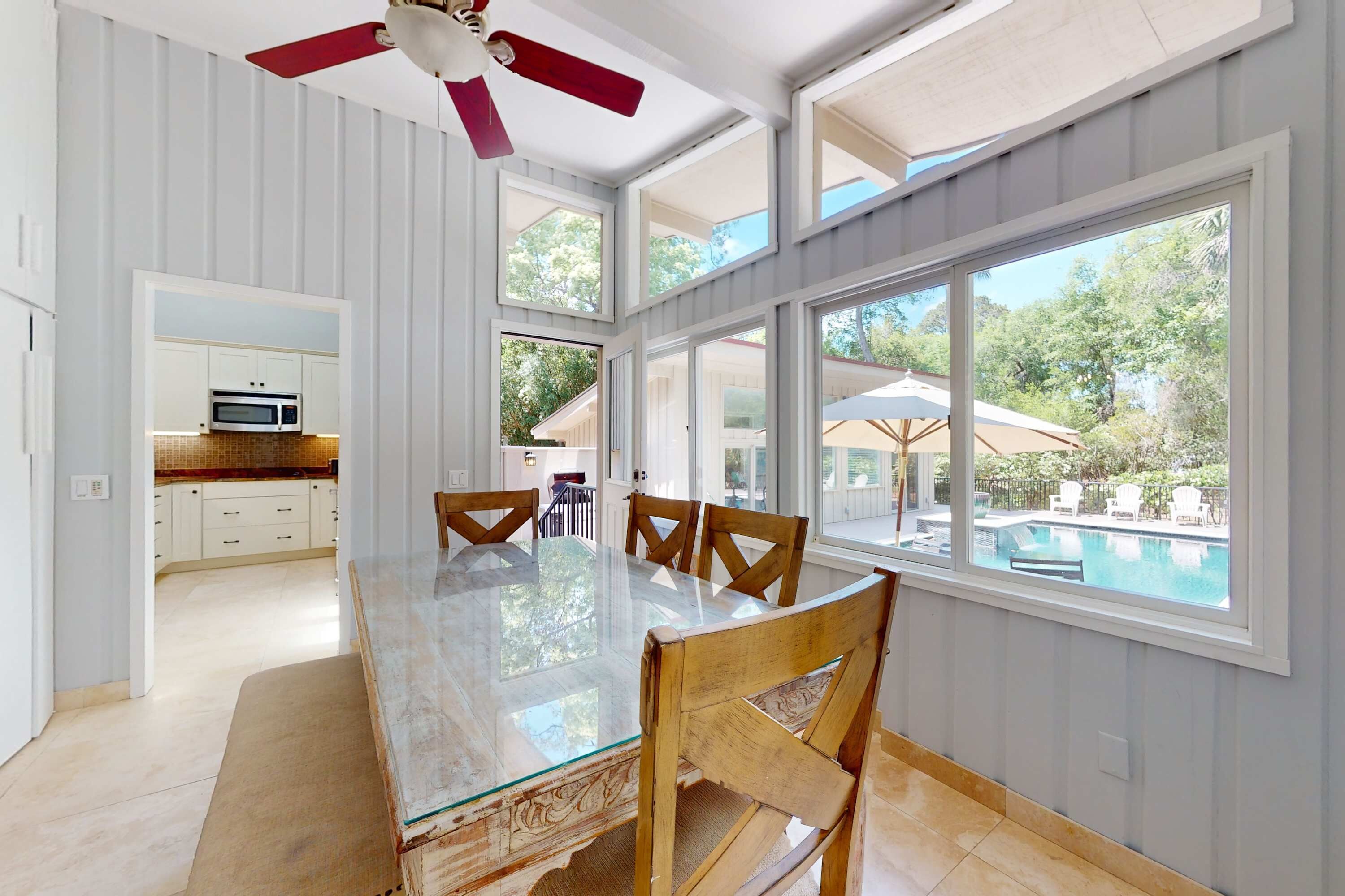 Uma área de refeições com mesa de vidro, cadeiras de madeira e grandes janelas com vista para uma piscina.