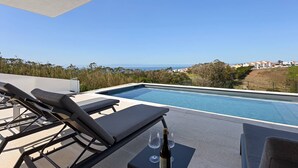 Ein Poolbereich mit Liegestühlen und einer Weinflasche, mit Blick auf einen klaren blauen Himmel und eine entfernte Häuseransicht.