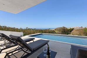 A poolside area with lounge chairs and a bottle of wine, overlooking a clear blue sky and a distant view of houses. - Villa - Casa Nunca Desista (Atalaia)