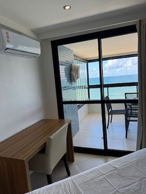 A room with a bed, a desk, and a view of the ocean through a sliding glass door. - Beachfront Apartment (Salvador)