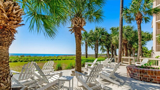 A deck with white Adirondack chairs, palm trees, and a view of the ocean. - Sunny condo with ocean view balcony & beach access (Hilton Head)