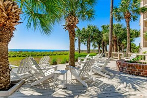 A deck with white Adirondack chairs, palm trees, and a view of the ocean. - Sunny condo with ocean view balcony & beach access (Hilton Head)