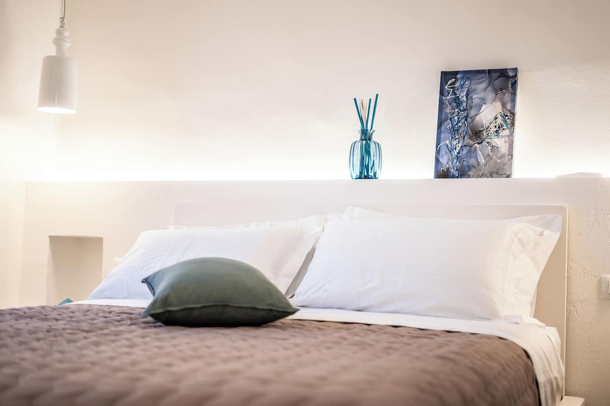 A neatly made bed with white linens and a green pillow, a blue vase with reeds, and a painting on the wall.
