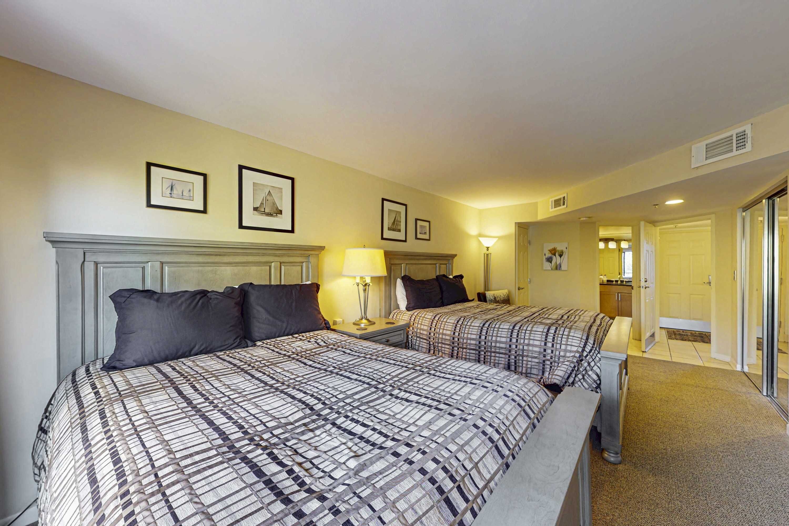 Une chambre d’hôtel avec deux lits, une table de chevet, une lampe, des tableaux encadrés au mur et une porte donnant sur une salle de bain.