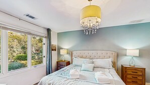 A bedroom with a large bed, two bedside tables, a chandelier, and a view of greenery through the window. - Fully Renovated Sea Pines Home w Private Pool Six Rows from the Water  Dogs OK (Hilton Head)