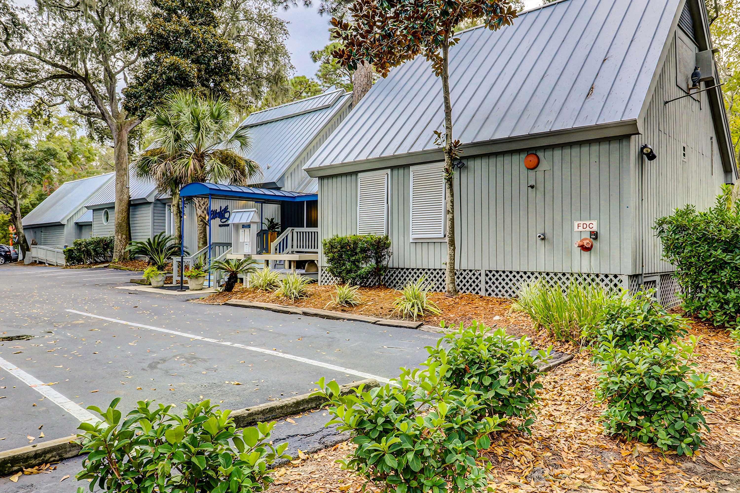 A row of single-story buildings with a covered outdoor seating area, a fenced parking lot, and a small garden area with shrubs and trees.