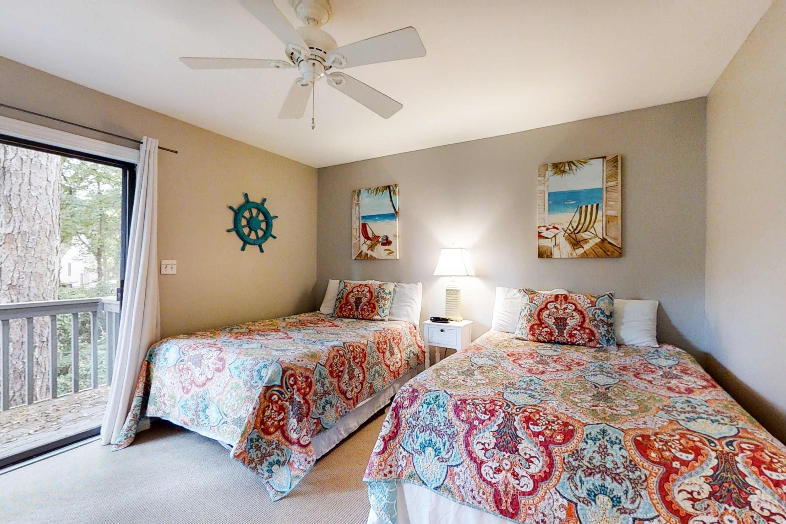 A room with two beds, a ceiling fan, a window with curtains, and artwork on the walls.