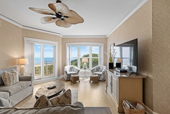 A spacious living room with a ceiling fan, a television, and a view of the outdoors through large windows. - Bright & Airy Retreat with Balcony Views, Walkable Beach Access & Central Location (Hilton Head)