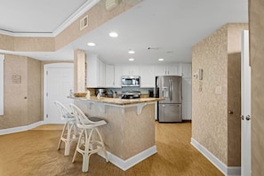 A kitchen with white cabinets, a stainless steel refrigerator, a microwave, and a coffee maker. There are two white bar stools with cushioned seats. - Bright & Airy Retreat with Balcony Views, Walkable Beach Access & Central Location (Hilton Head)