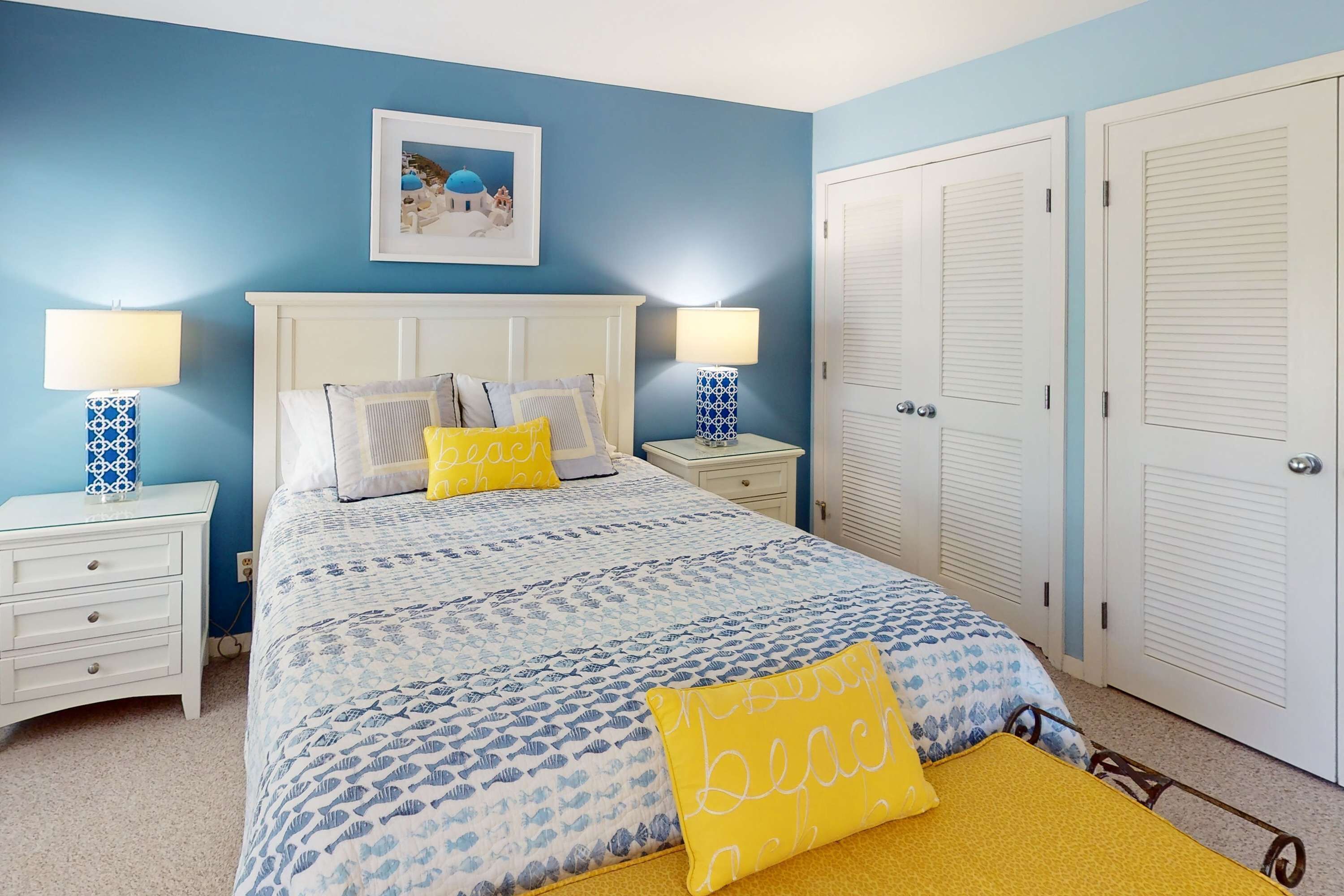 Une chambre à coucher comprenant un lit, deux tables de chevet, une commode, un tableau accroché au mur et un ensemble de portes blanches.
