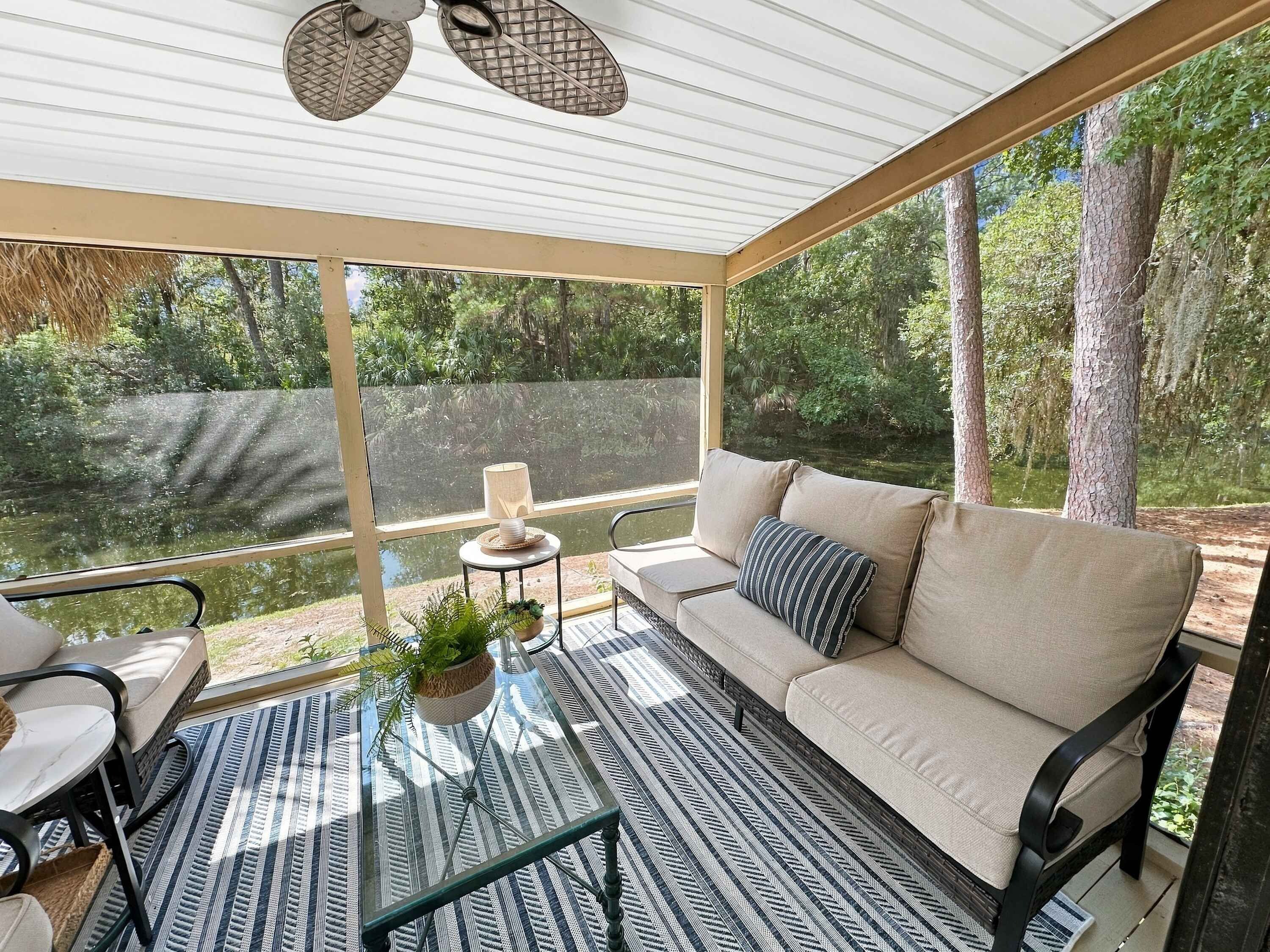 A covered outdoor seating area with a sofa, chairs, and a table, overlooking a body of water and surrounded by trees.