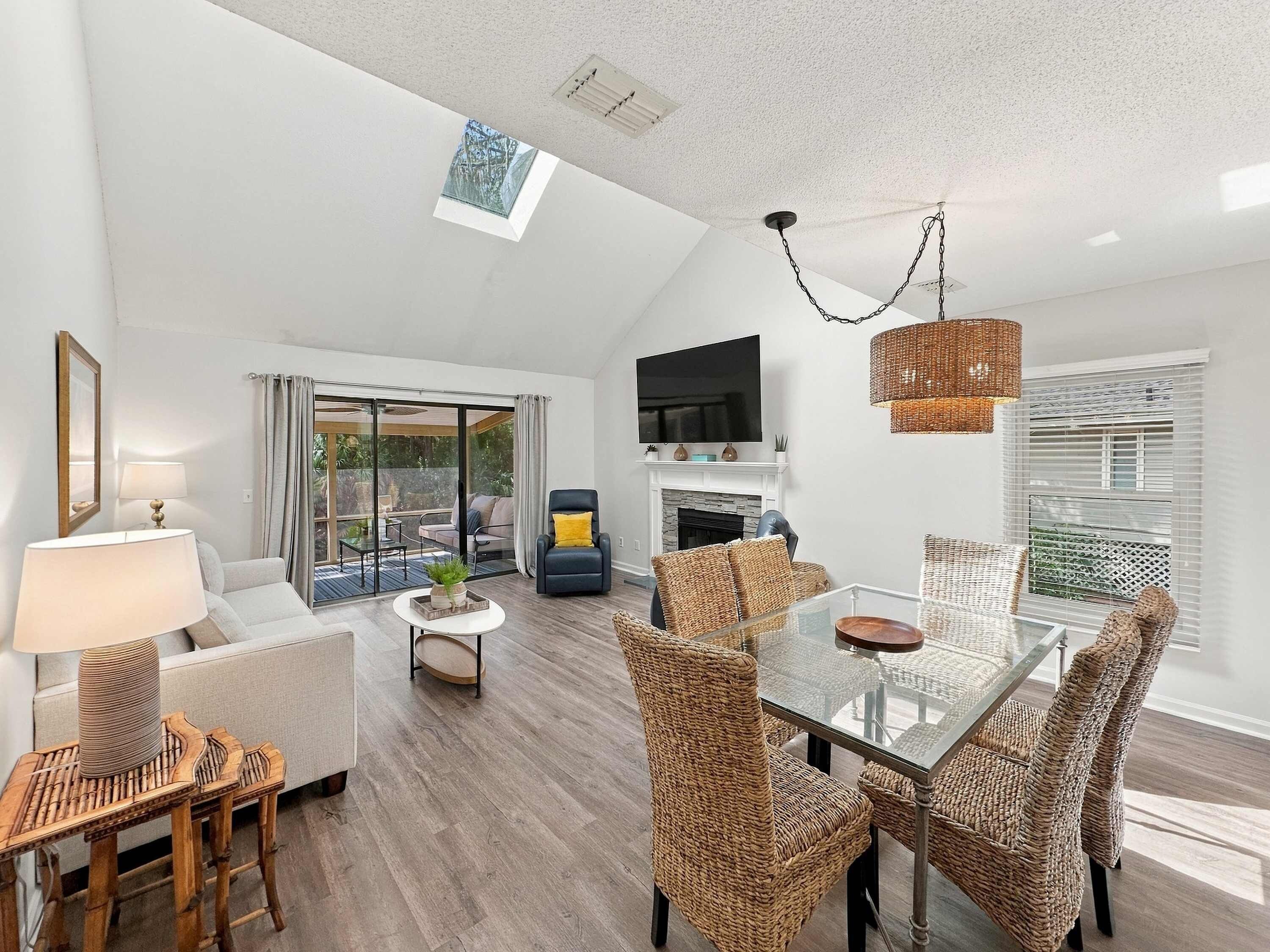 A modern living room with a glass dining table, wicker chairs, a flat-screen TV, and a fireplace.