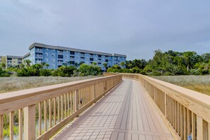 A wooden boardwalk leading to a blue building with multiple balconies, surrounded by greenery. - Charming Coastal Villa with Pools, Hot Tub, Gym & Private Beach Access (Hilton Head)