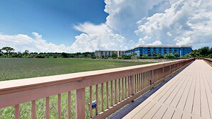 A wooden boardwalk leading to a building with a blue facade, surrounded by a grassy field and trees. - Oceanside Resort Condo with Indoor Pool, Tennis & Oceanfront Restaurant (Hilton Head)
