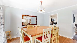 Un comedor con una mesa y sillas de madera, una araña y una cocina visible a través de un vano abierto.