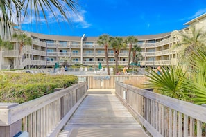 A multi-story hotel with balconies, a pool area, and a wooden boardwalk leading to the entrance. - Beach-view condo in oceanfront complex with outdoor pool, beach access & balcony (Hilton Head)
