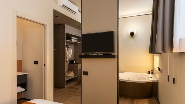 A modern hotel room with a bed, a television, a shower area, and a walk-in closet. - Relax Hot Tub – San Giovanni (Rome)