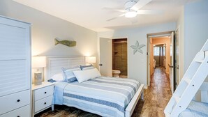 A bedroom with a bed, nightstands, a ceiling fan, a wooden floor, a bathroom with a toilet, and a staircase.