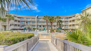Un hotel de varios pisos con balcones, una zona de piscina y un paseo marítimo de madera que conduce a la entrada.