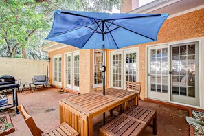 Eine Terrasse im Freien mit einem Holztisch, Bänken, einem blauen Sonnenschirm und einem Grill.
