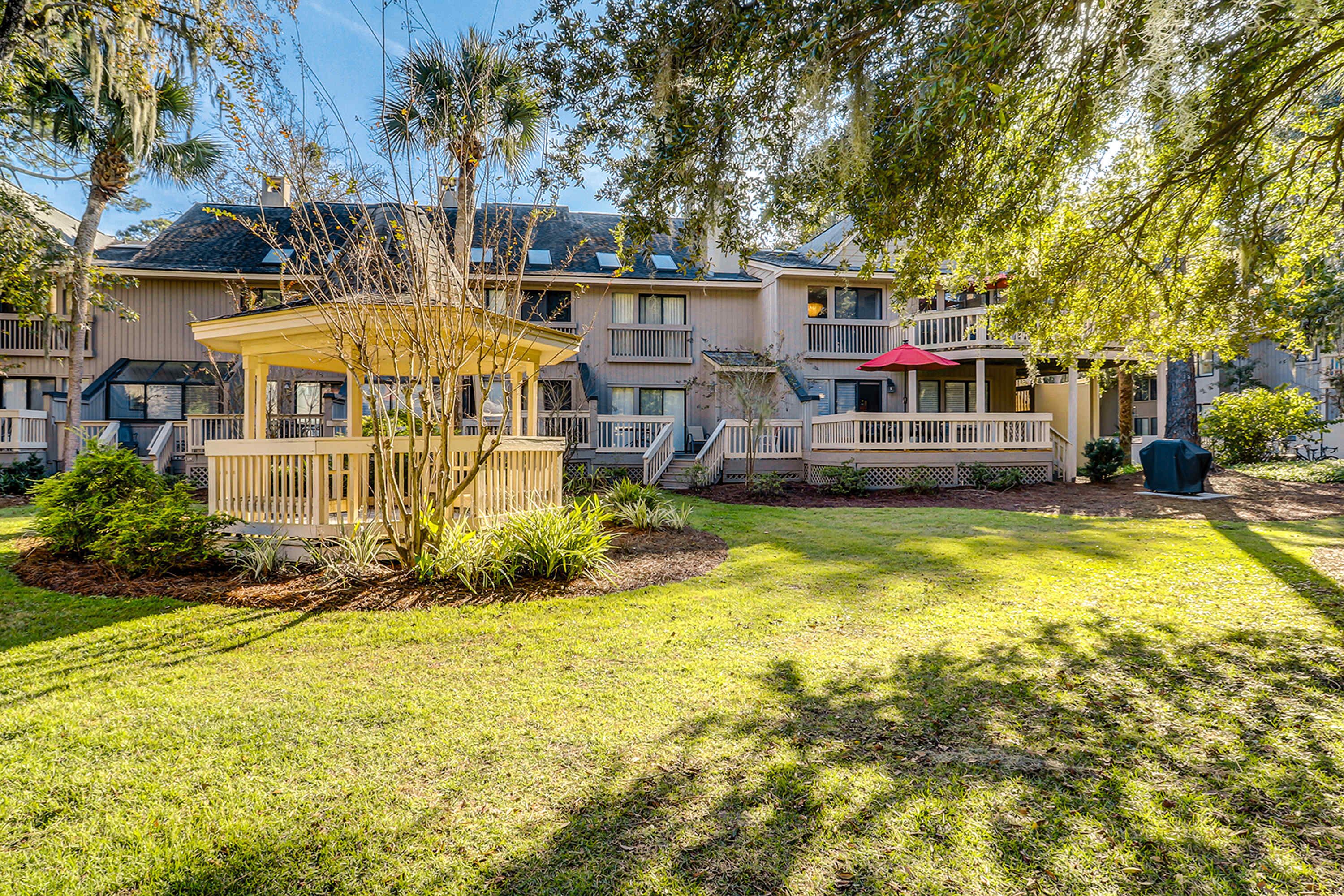 A multi-story hotel with a covered outdoor seating area, a well-maintained lawn, and a variety of trees.