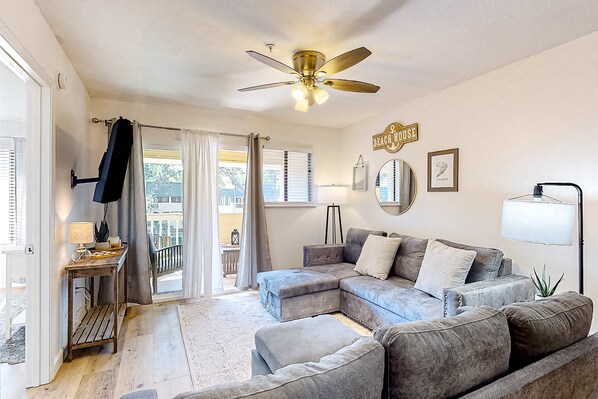 Ein Wohnzimmer mit einer Eckcouch, einem Deckenventilator, einem kleinen Tisch mit Lampe und einem Schild mit der Aufschrift 'Beach House'.
