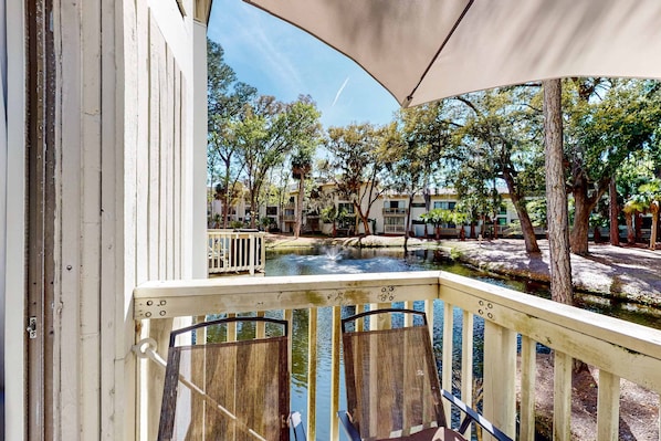 A balcony with wooden chairs, a small pond, and trees in the background. - Hilton Head Island condo with water-view patio, pool, & easy beach access (Hilton Head)