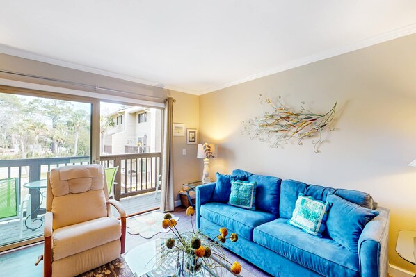 A living room with a blue sofa, a beige armchair, a glass coffee table, and a view of the outside. - Fun & bright beach getaway with pool, easy beach access & pond view (Hilton Head)